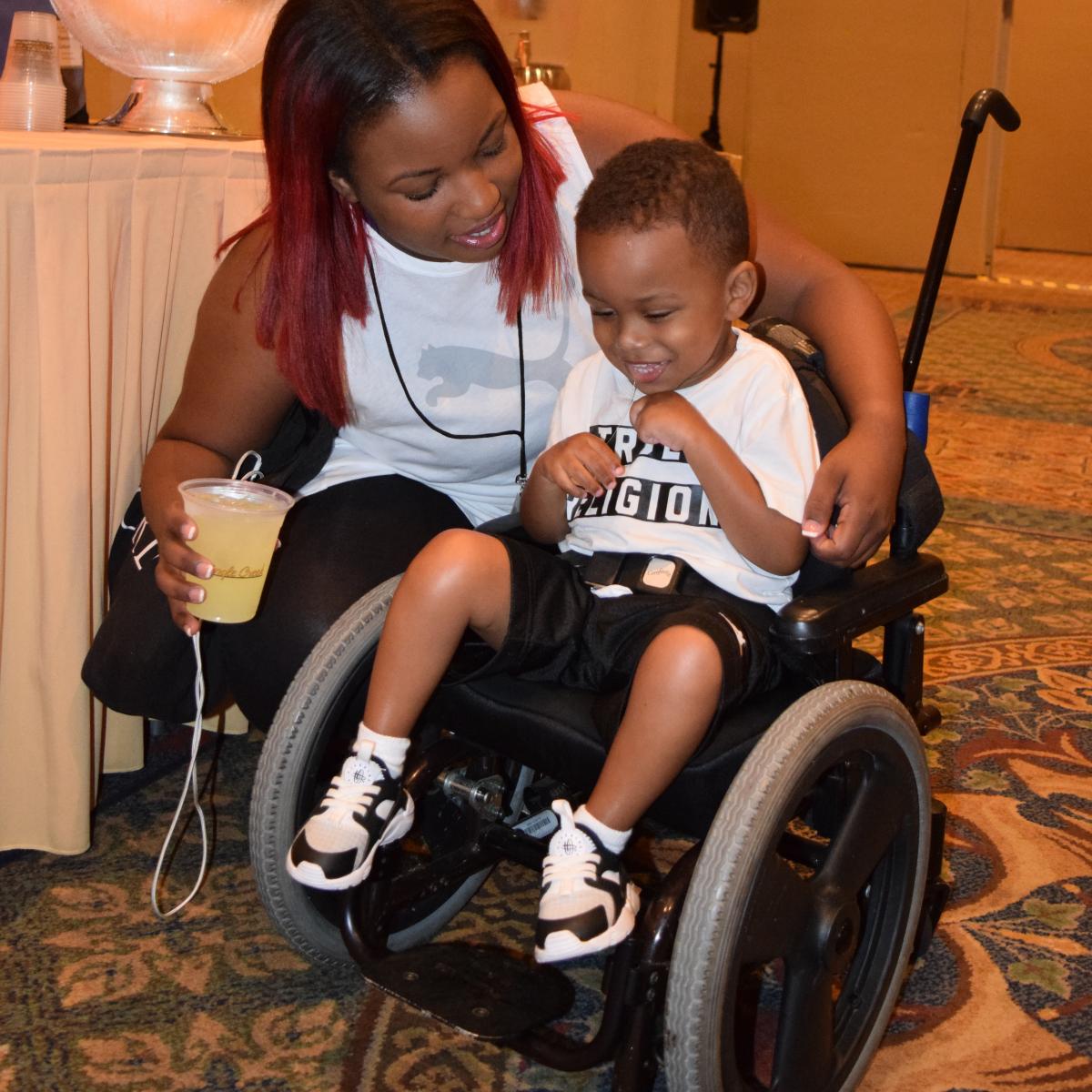 a small child smiling in his wheelchair with his mother