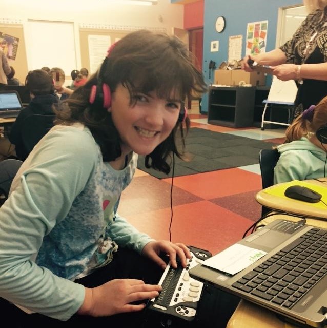 a girl in a classroom wearing headphone at a laptop using a braille display, She is smiling